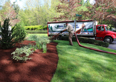 A Bark King blower truck at the kerb with a crew member running hose up a planted bed